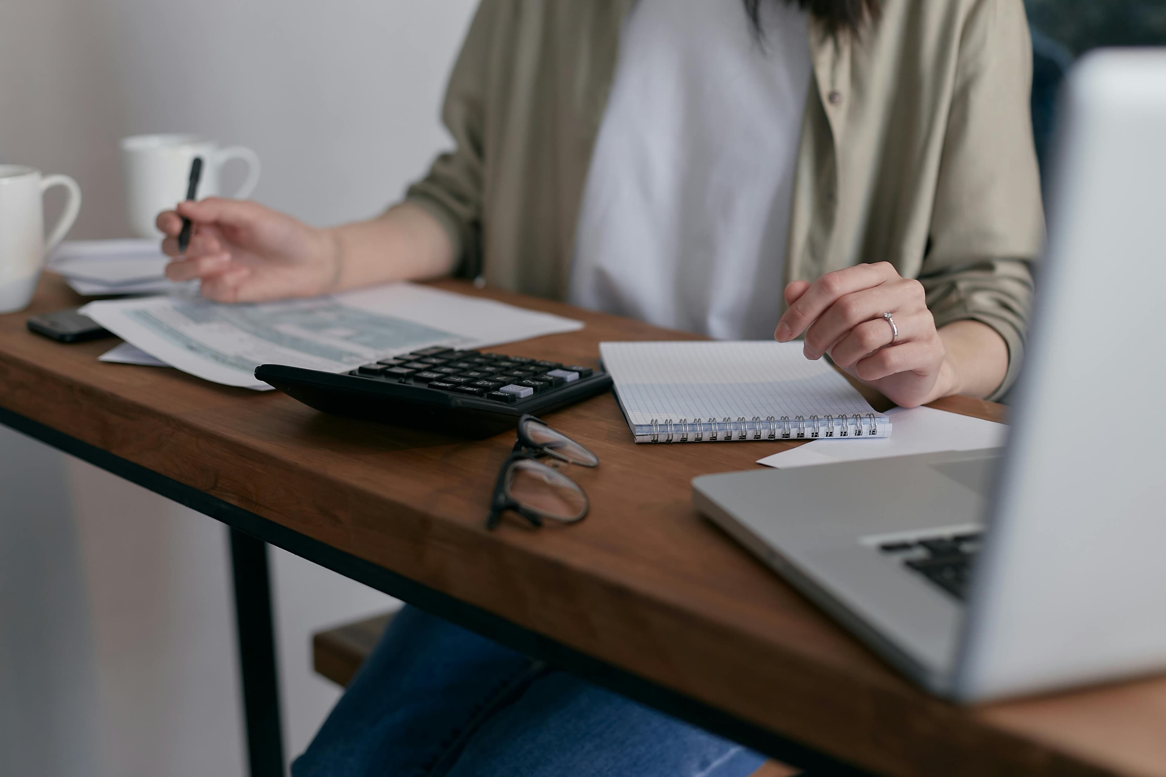 Person working at a wooden desk with a calculator, papers, and a laptop, taking notes with a pen and notebook.