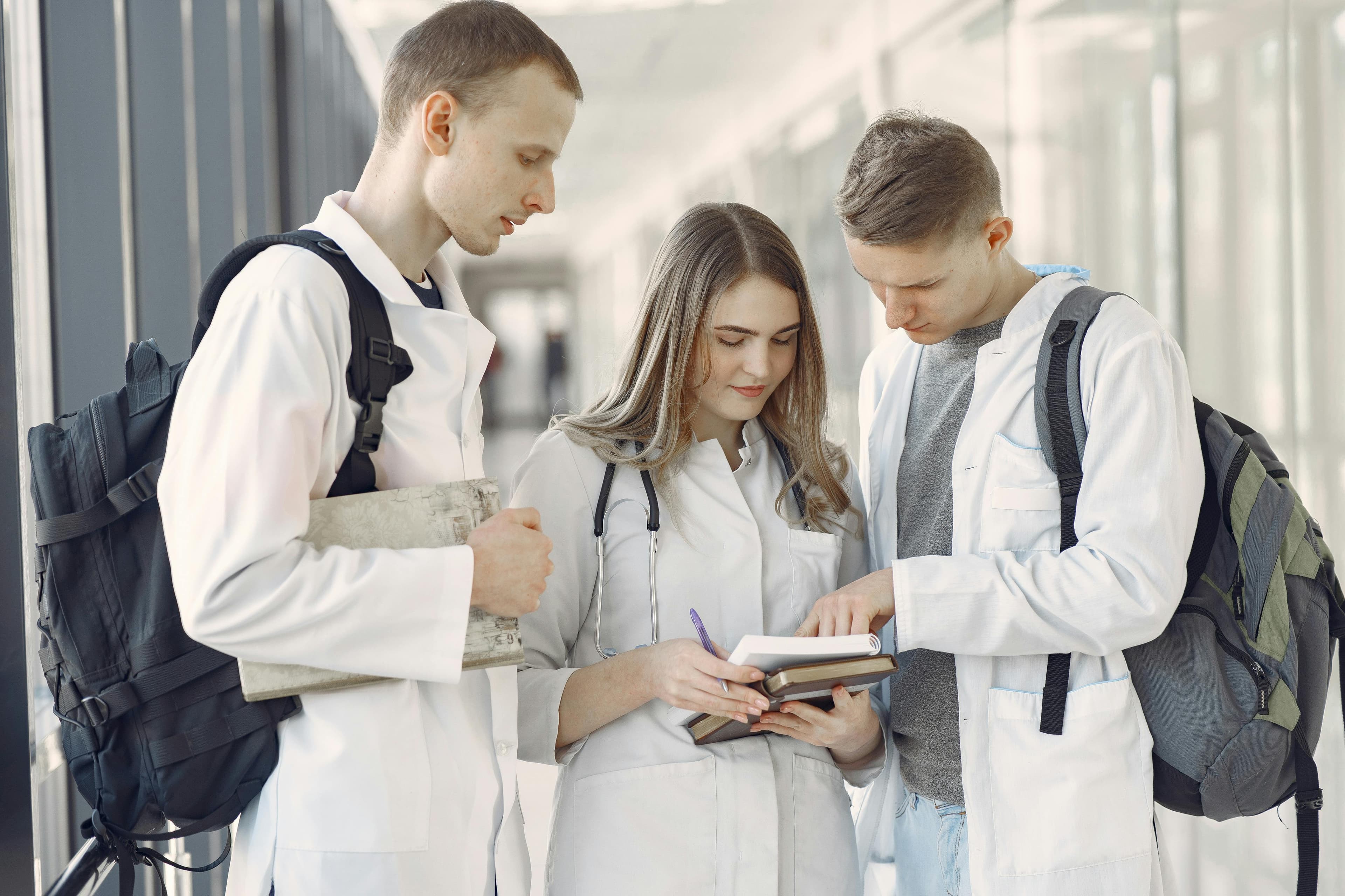 Group of three medical students in white coats standing in a hallway, reviewing notes together.