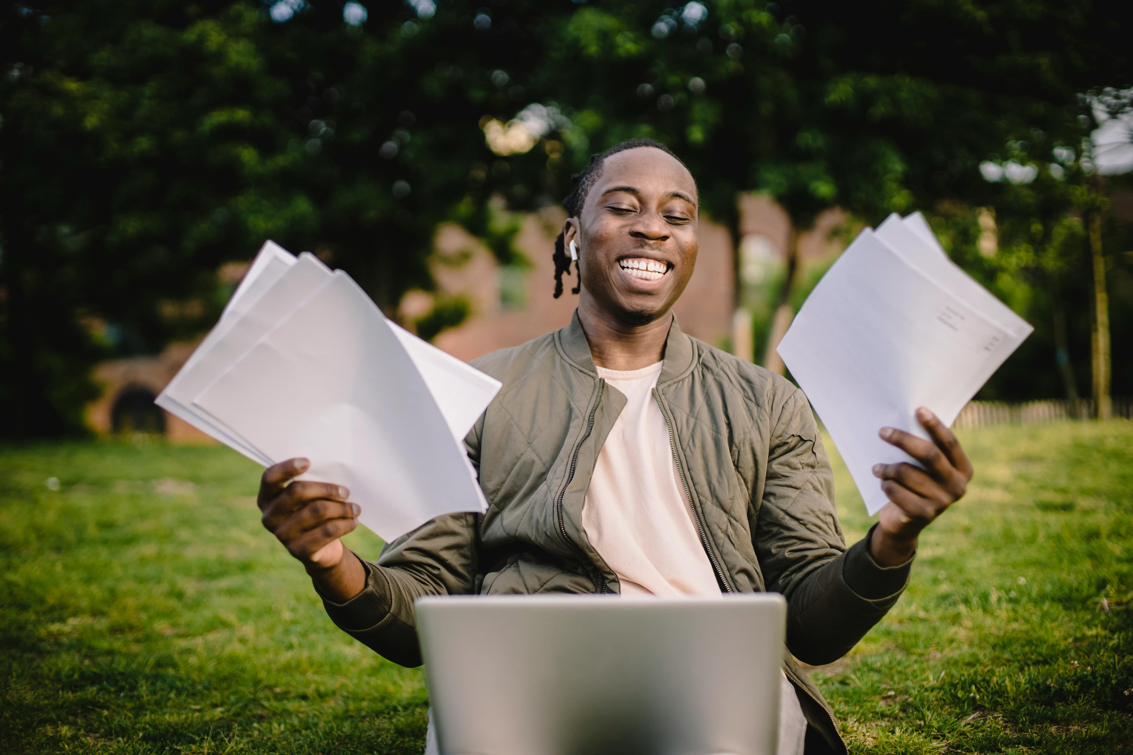 Smiling student sitting outdoors with a laptop, holding up sheets of paper in both hands.