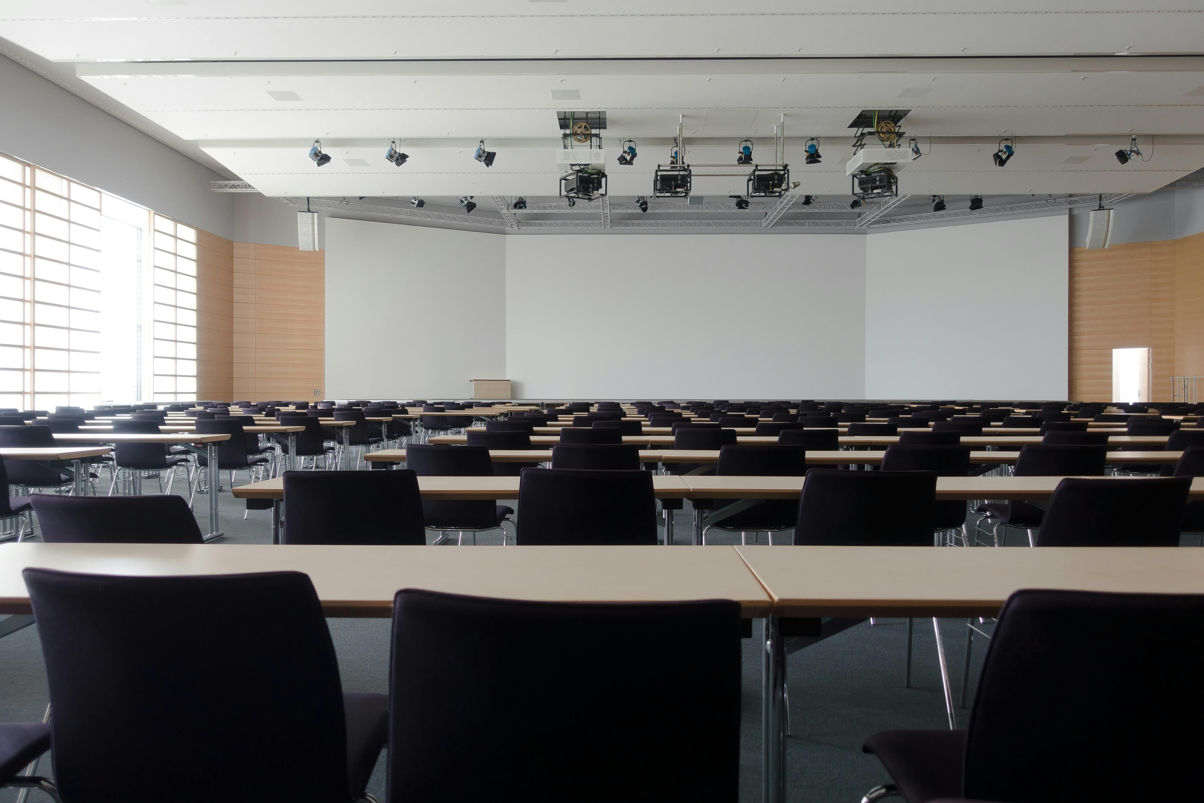 Empty lecture hall with rows of desks and chairs facing a large white presentation screen.