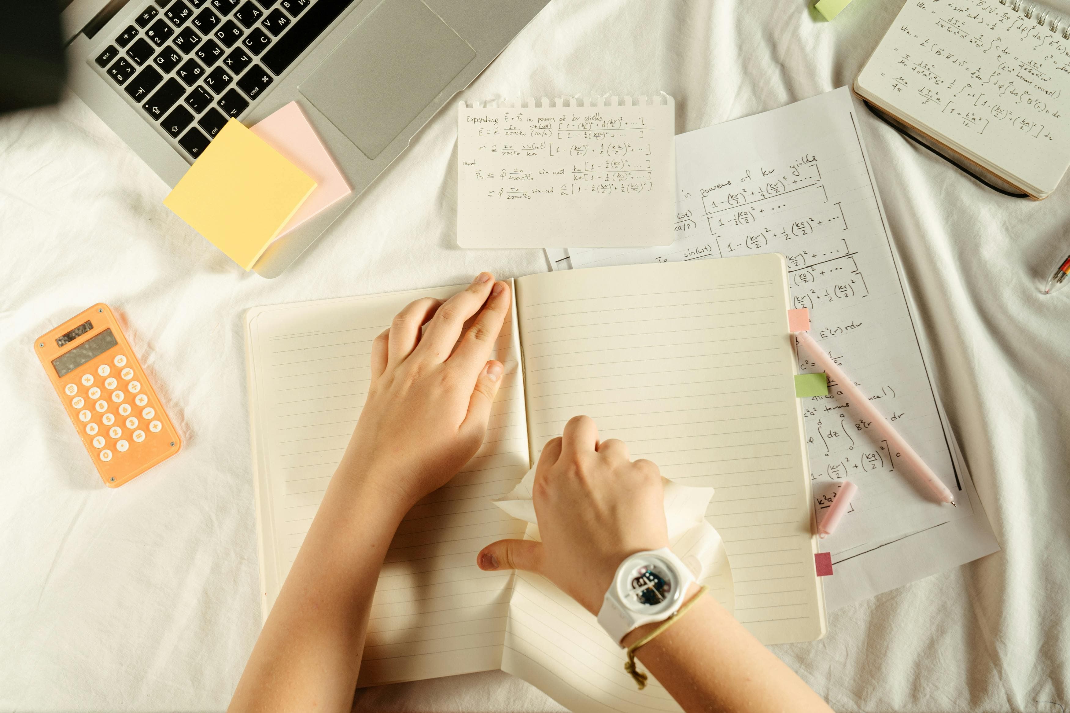 Student studying at home with open notebook, laptop, calculator, and handwritten math notes on the bed.
