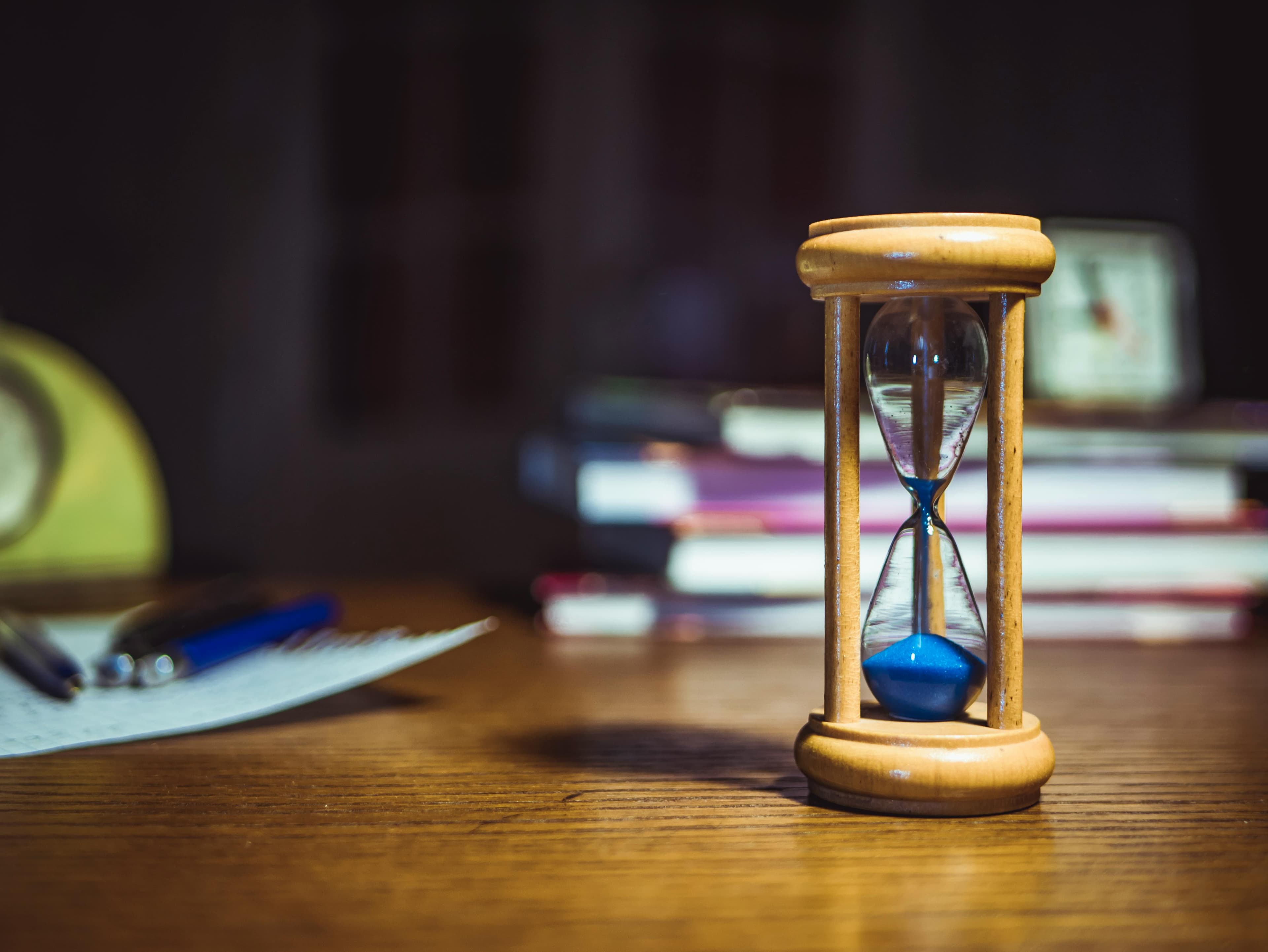 Wooden hourglass with blue sand on a desk beside papers, pens, and stacked books.