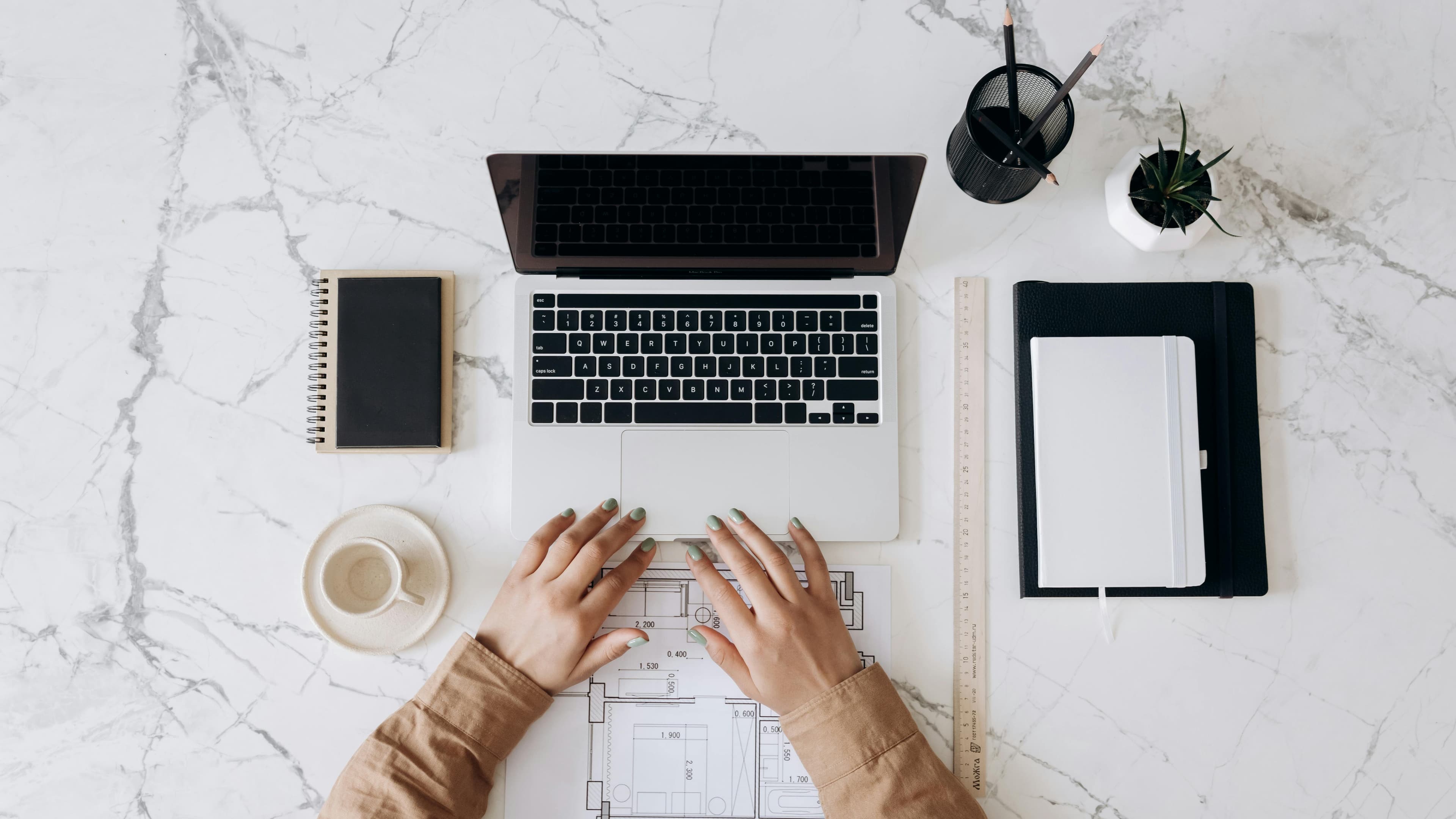 Person working on a laptop at a marble desk with notebooks, a ruler, pencils, and a cup of coffee.